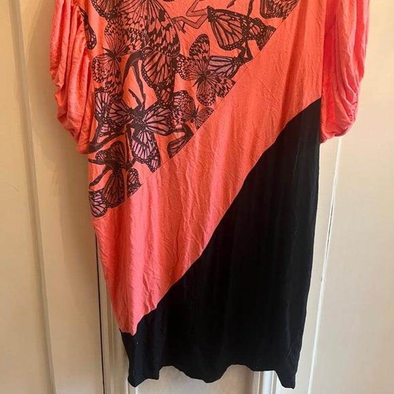 May include: Coral and black asymmetrical top. The coral section features a butterfly pattern in shades of brown and black. The top has short sleeves and a diagonal color block design.