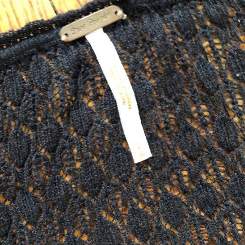 Pu&ograve; includere: Primo piano di un capo nero a trama aperta con un motivo a rombi. Un'etichetta color oro con la scritta "Free People" &egrave; attaccata. &Egrave; attaccata anche un'etichetta bianca con testo.