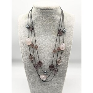 Op de afbeelding: Een meerstrengs ketting met roze en grijze kralen. De ketting is gemaakt van zwart koord en heeft een verscheidenheid aan ronde en ovale kralen.