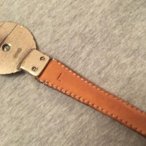 Op de afbeelding: Close-up van een bruinleren riem met een zilveren gesp. De riem heeft een gestikte rand en de letter "L" is zichtbaar op het leer. De gesp is rond en heeft een metalen afwerking.