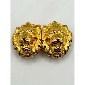 May include: Two gold-colored lion head embellishments. Each lion head is highly detailed with a textured mane, facial features, and a slightly open mouth. The embellishments are likely made of metal and have a shiny, reflective surface.