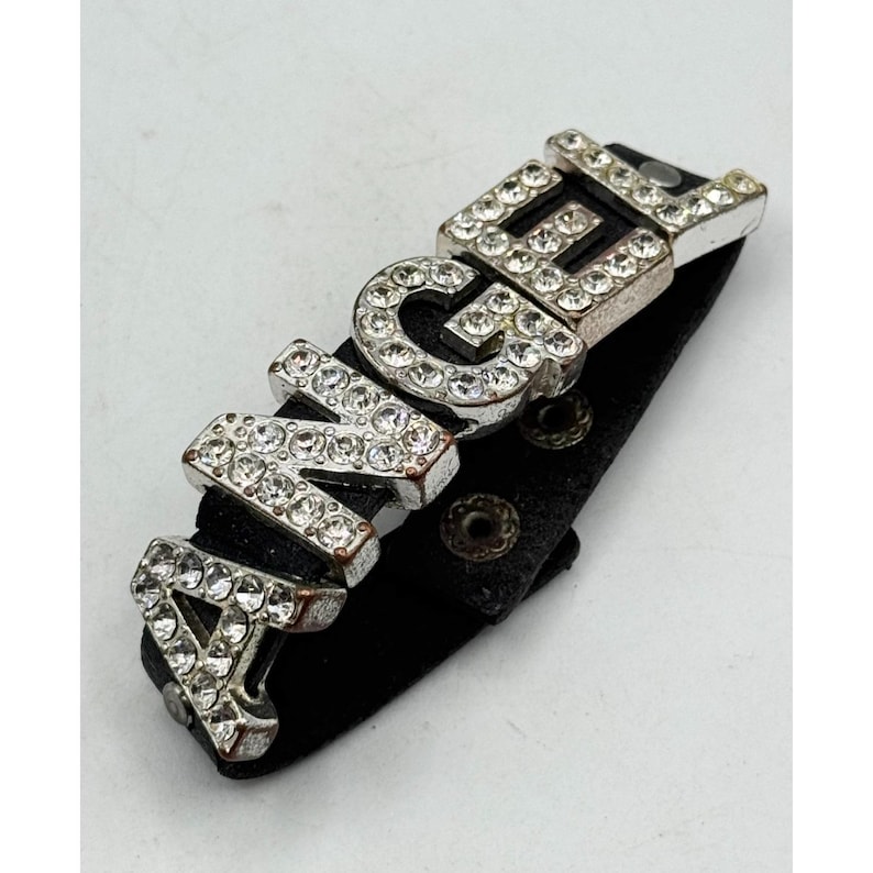 May include: A black leather bracelet with the word "ANGEL" spelled out in silver-toned metal letters encrusted with clear rhinestones. The bracelet has a snap closure.
