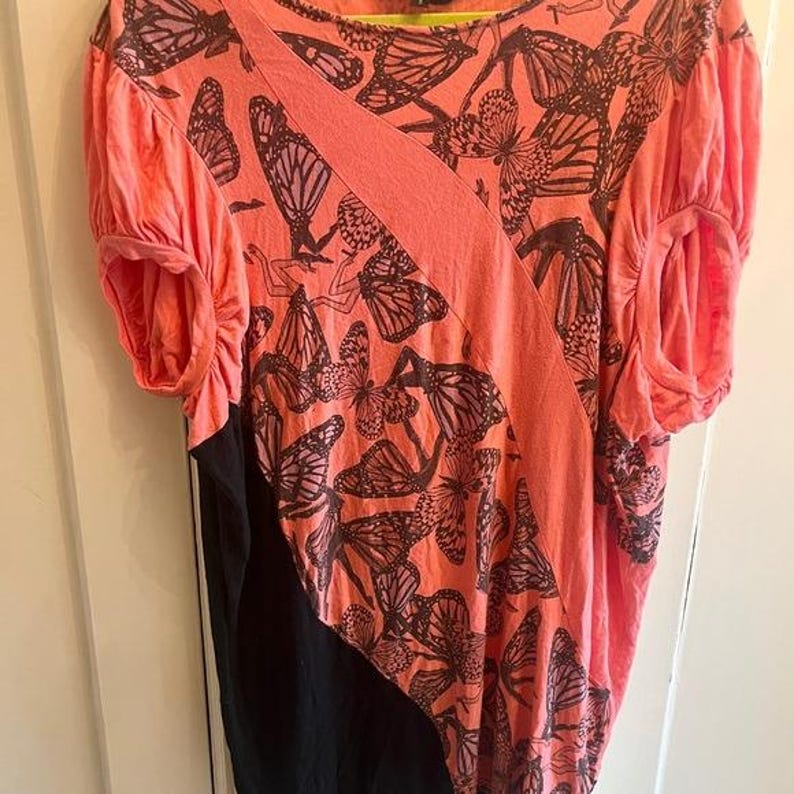 May include: Coral-colored top with a butterfly print and black panel. The short-sleeved shirt features a diagonal design across the front and gathered sleeves. The butterfly pattern is in shades of brown and black.