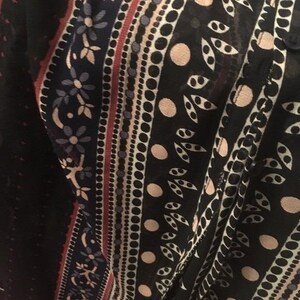 Op de afbeelding: Close-up van een zwart kledingstuk met patroon. Het ontwerp heeft verticale strepen met bloemen-, stip- en bladpatronen in tinten wit, rood en beige. De stof lijkt licht en semi-transparant.