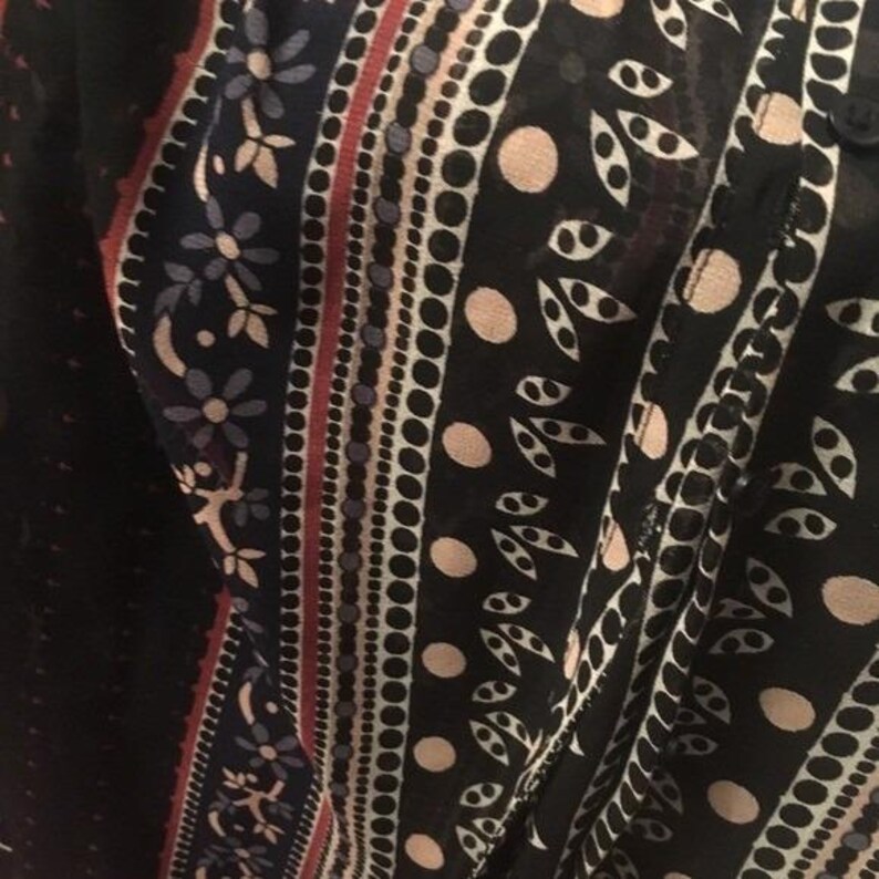 Op de afbeelding: Close-up van een zwart kledingstuk met patroon. Het ontwerp heeft verticale strepen met bloemen-, stip- en bladpatronen in tinten wit, rood en beige. De stof lijkt licht en semi-transparant.