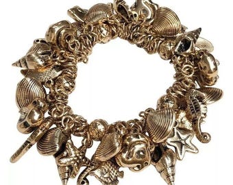 May include: A gold-toned charm bracelet featuring a variety of seashell and starfish charms. The bracelet is made of a chain link design and is perfect for adding a touch of summer to any outfit.