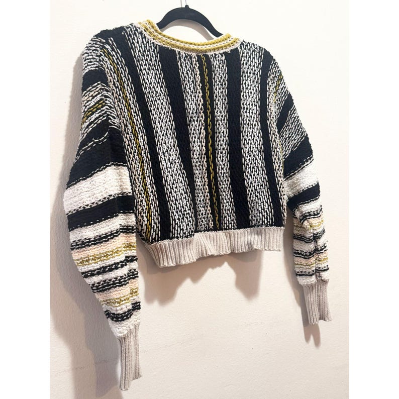 Puede incluir: Un su&eacute;ter a rayas corto con mangas largas. El su&eacute;ter es negro, blanco y amarillo. Las mangas tienen un patr&oacute;n de rayas multicolores.