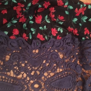 Puede incluir: Primer plano de un textil con estampado floral y ribete de encaje. La parte superior presenta una tela negra con flores rojas y hojas verdes. La secci&oacute;n inferior es de encaje azul marino con dise&ntilde;os intrincados.