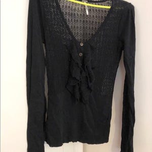 Pu&ograve; includere: Camicia nera a maniche lunghe con scollo a V e parte anteriore a balze. La parte superiore del petto presenta un motivo a maglia aperta e si chiude con due bottoni. Le maniche sono lunghe e aderenti.