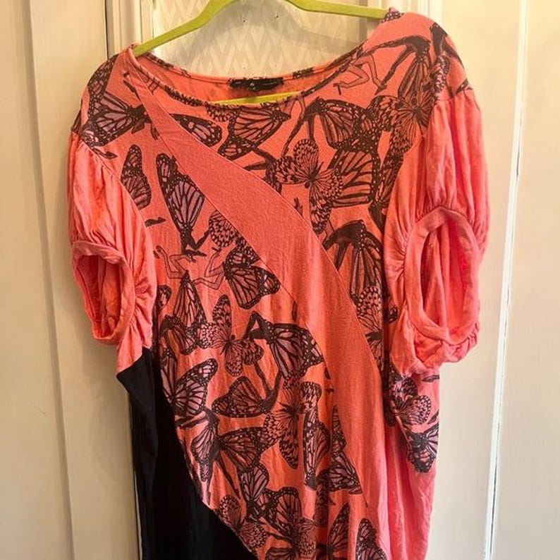 May include: Coral-colored top with a butterfly print. The short-sleeved shirt features a diagonal coral stripe across the front and a black panel on the bottom. The butterfly pattern is in a dark brown color.