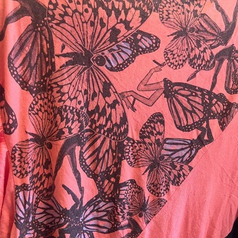 May include: Coral-colored t-shirt featuring a repeating pattern of large butterflies in shades of black and purple. The butterfly wings have intricate details. The design also includes a stylized illustration of a person.