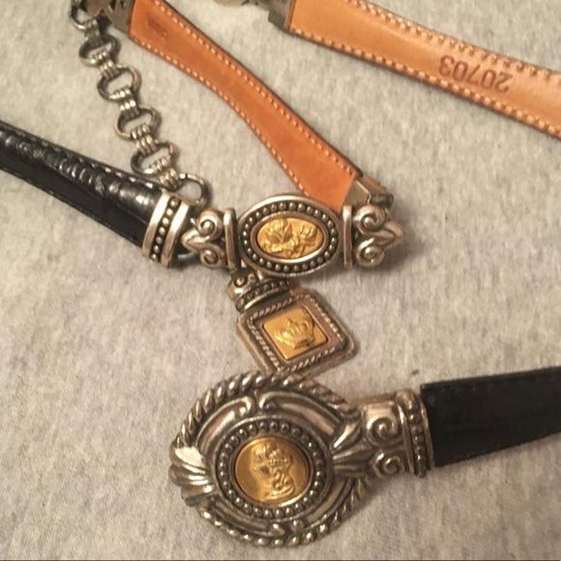 Op de afbeelding: Close-up van twee riemen met sierlijke zilveren gespen en gouden accenten. E&eacute;n riem is zwart, de andere is bruin leer. De gespen hebben reli&euml;fontwerpen en een kroonbedel.