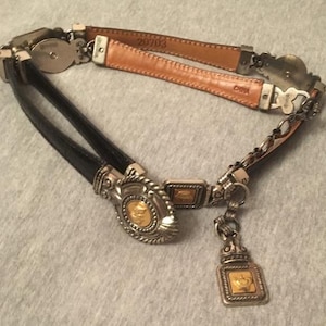 Op de afbeelding: Een decoratieve riem met een zwarte leren band en een bruine leren band. De riem heeft zilverkleurige metalen accenten en een goudkleurig medaillon met een kroonontwerp. De riem is ontworpen met een ketting en een bungelende bedel.