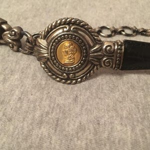 Op de afbeelding: Zilverkleurige riem met een zwarte leren band. De gesp heeft een goudkleurig medaillon met een profielportret, omgeven door sierlijke zilveren details en een ketting. Een stijlvol accessoire.