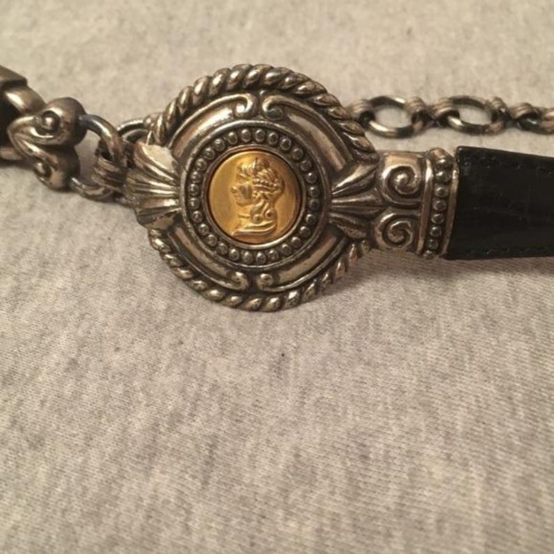 Op de afbeelding: Zilverkleurige riem met een zwarte leren band. De gesp heeft een goudkleurig medaillon met een profielportret, omgeven door sierlijke zilveren details en een ketting. Een stijlvol accessoire.