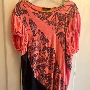 May include: Coral-colored top with a butterfly pattern. The short-sleeved shirt features a diagonal coral stripe and a black panel on the lower left side. The butterfly design is in shades of brown and black.