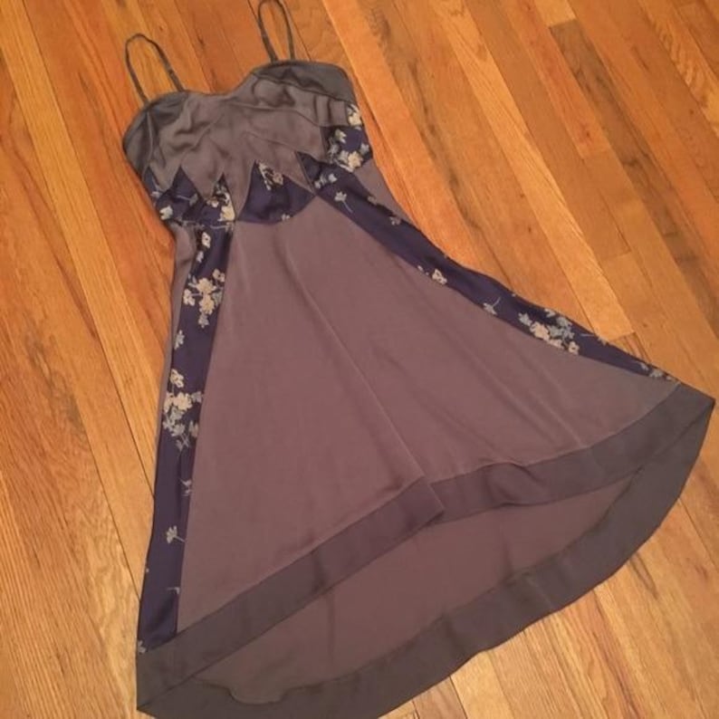 May include: A sleeveless dress with spaghetti straps. The dress features a dark gray bodice and a flowing skirt in a muted purple hue. Dark blue panels with a floral pattern accent the dress.