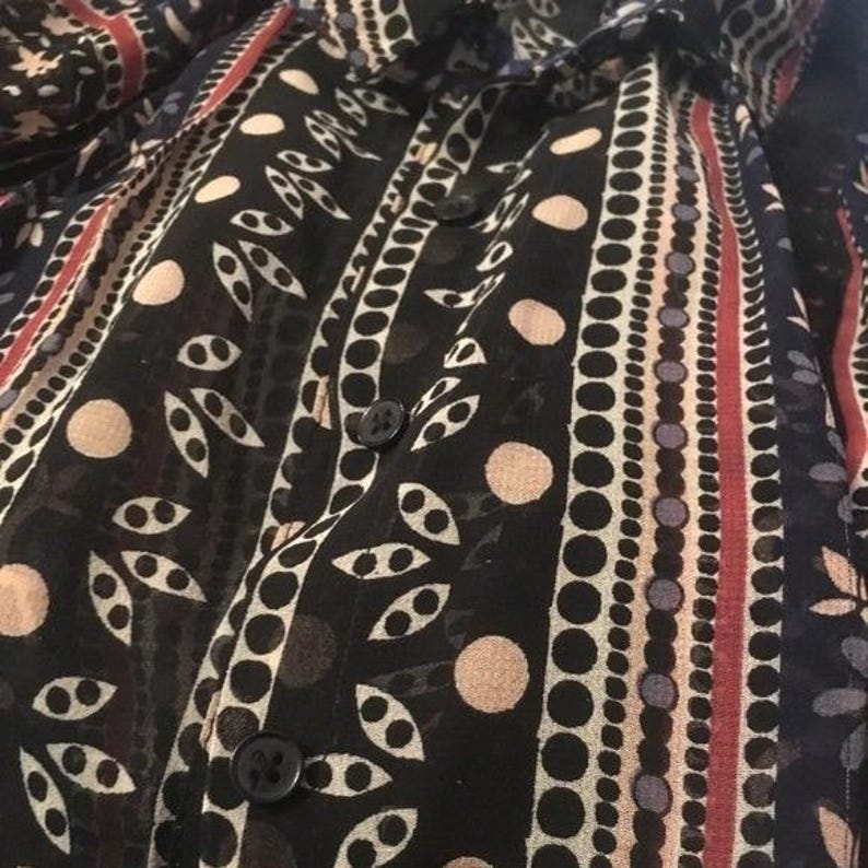 Op de afbeelding: Close-up van een zwart overhemd met knopen en een semi-transparante stof. Het overhemd heeft verticale strepen met afwisselende patronen van stippen, bladeren en lijnen. Het kleurenpalet omvat zwart, wit, rood en beige.