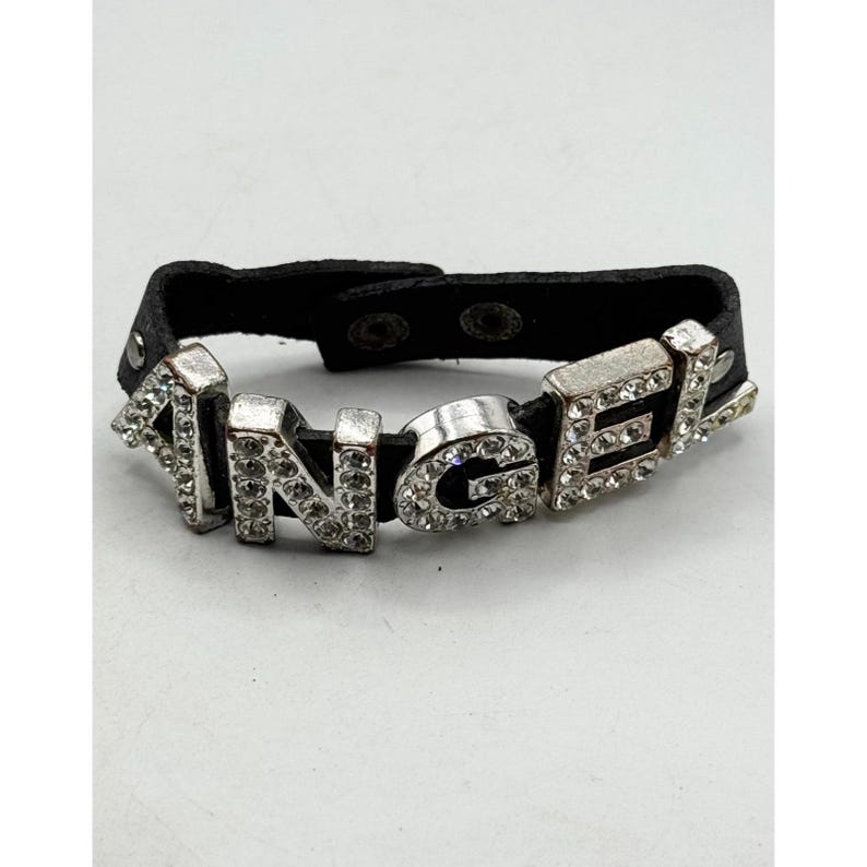 May include: A black leather bracelet with the word "ANGEL" spelled out in silver letters. Each letter is encrusted with small, clear rhinestones. The bracelet has a snap closure.