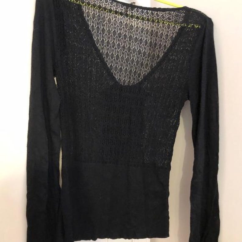 Pu&ograve; includere: Top nero a maniche lunghe con scollo a V e un esclusivo design a maglia aperta sulla parte superiore del petto e sulla schiena. La parte inferiore del top &egrave; in tessuto nero tinta unita. Il top &egrave; appeso a una gruccia gialla.