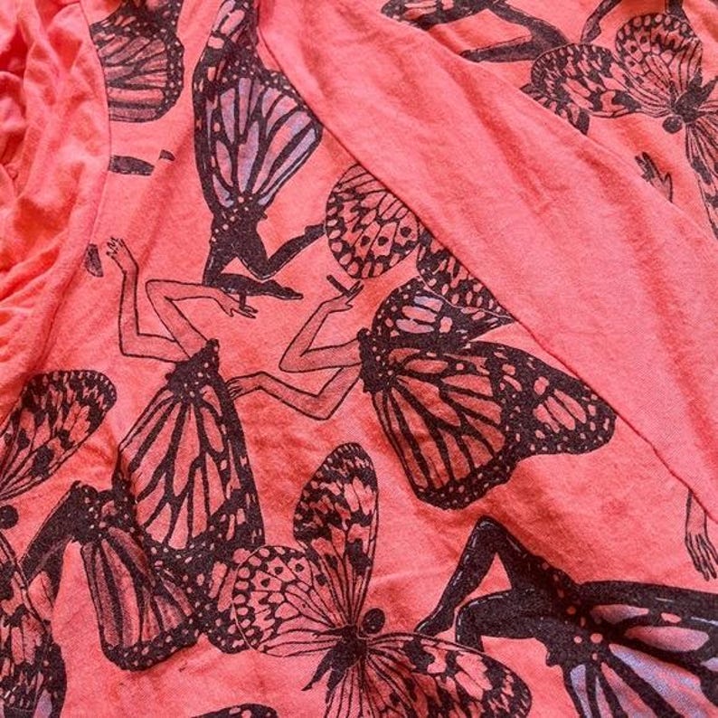 May include: Coral-colored fabric with a repeating pattern of black butterfly figures with human-like bodies. The butterflies have detailed wing patterns and are in various poses. The fabric is slightly wrinkled, suggesting it is clothing.