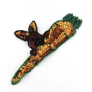 Könnte beinhalten: Eine braune und goldene Pailletten-Hasenapplikation, die eine grüne und goldene Pailletten-Karotte hält.