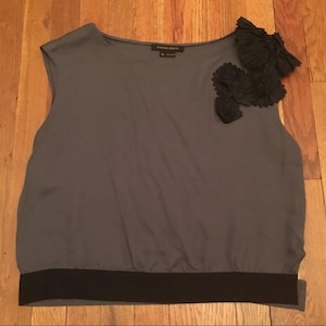 Puede incluir: Una blusa sin mangas gris con una banda negra en la parte inferior y adornos florales negros en el hombro. La blusa está hecha de una tela suave y fluida.