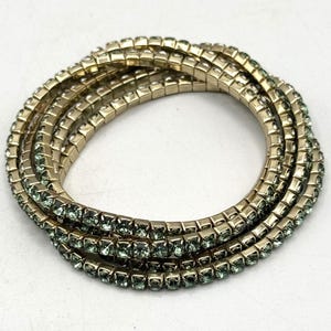 May include: A set of five gold-toned bracelets with light green rhinestones. The bracelets are layered on top of each other.