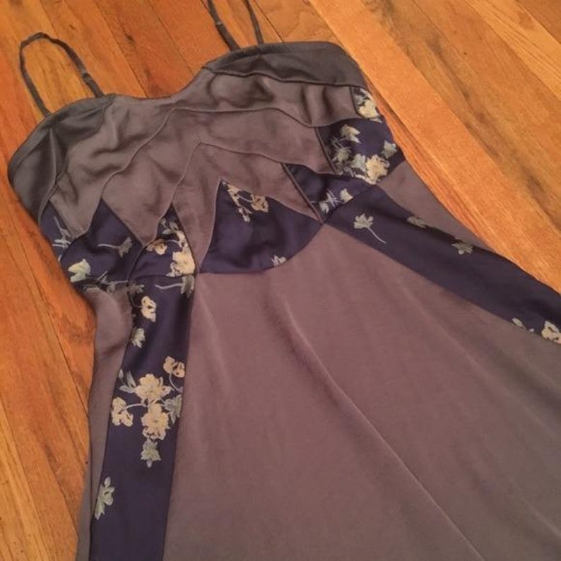 May include: A sleeveless, floor-length dress in shades of gray and purple. The bodice features layered panels and floral patterned accents in navy blue. The dress has thin spaghetti straps.