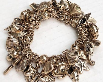 May include: A gold-toned charm bracelet featuring various sea-themed charms, including seashells, starfish, and seahorses. The bracelet is made of linked chains and has a circular design. The charms are detailed and add a nautical touch.