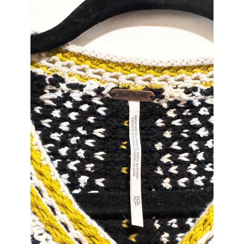 Puede incluir: Su&eacute;ter de crochet negro, blanco y amarillo con una etiqueta de Free People. La etiqueta dice "Knit in Taiwan, Finished in China" y "Tricote dans Taiwan, Fini en Chine".