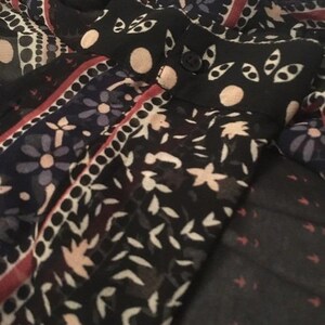 Op de afbeelding: Close-up van een patroonstof met een zwarte achtergrond. De stof heeft bloemen en geometrische ontwerpen in wit, rood en blauw. De kraag heeft een knoop en is zwart met witte stippen en bladachtige patronen.