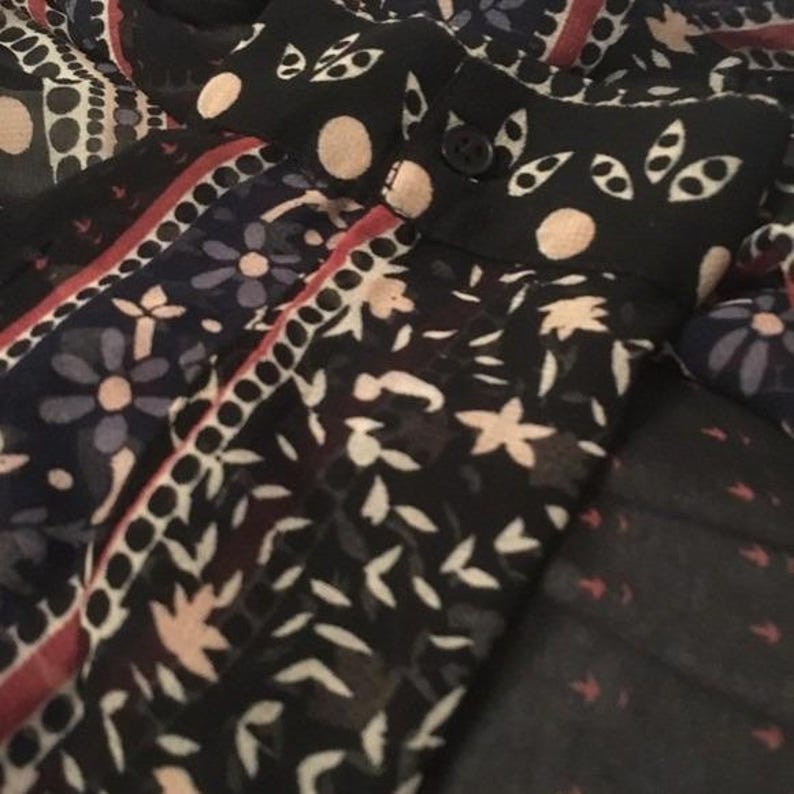 Op de afbeelding: Close-up van een patroonstof met een zwarte achtergrond. De stof heeft bloemen en geometrische ontwerpen in wit, rood en blauw. De kraag heeft een knoop en is zwart met witte stippen en bladachtige patronen.