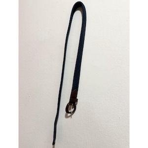 Op de afbeelding: Een marineblauwe gevlochten riem met een bruine leren gesp. De riem is lang en dun, geschikt voor broeken of rokken. De gesp heeft een eenvoudig, rond ontwerp.