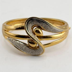 May include: A gold and silver tone bangle bracelet with a textured, abstract design. The bracelet is made of metal and has a hinged closure.