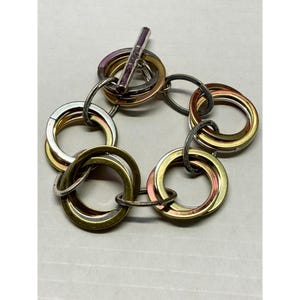 Puede incluir: Un brazalete de plata, oro y cobre con múltiples anillos entrelazados. El brazalete tiene un cierre de palanca.