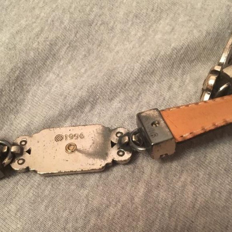 Op de afbeelding: Close-up van een zilverkleurige riem met een bruinleren band. De riem heeft decoratieve metalen schakels en een rechthoekige gesp met het jaartal "1996" erop gestempeld. De riem ligt op een grijze ondergrond.