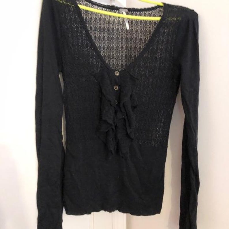 Pu&ograve; includere: Un maglione nero a maniche lunghe con scollo a V e parte anteriore a balze. Il maglione ha una chiusura con bottoni e un delicato motivo a maglia aperta sulla parte superiore del petto. L'indumento &egrave; appeso a una gruccia gialla.