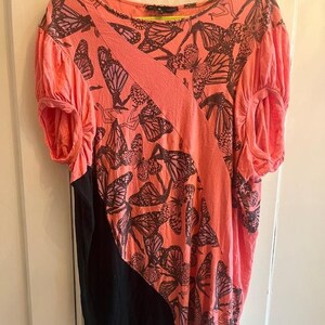 May include: Coral-colored top with a black panel and butterfly print. The short-sleeved shirt features a diagonal coral stripe across the front and a round neckline. The butterfly pattern is in dark brown.