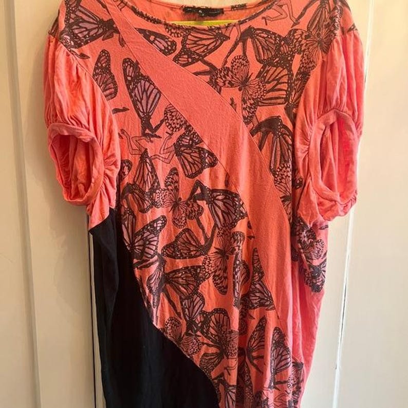 May include: Coral-colored top with a black panel and butterfly print. The short-sleeved shirt features a diagonal coral stripe across the front and a round neckline. The butterfly pattern is in dark brown.