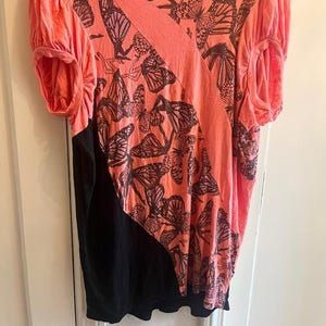 May include: A coral-colored top with a black lower section. The top features a butterfly print in dark brown and a diagonal design across the front. The sleeves are short and gathered, creating a puffy effect. The garment is made of soft fabric.