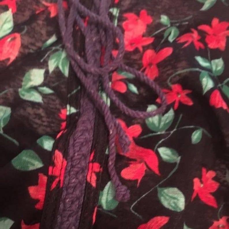 Puede incluir: Primer plano de una tela floral con flores rojas y hojas verdes sobre un fondo negro. Se aprecia un detalle de cuerda morada. La tela parece ligera, posiblemente para una blusa o un vestido.