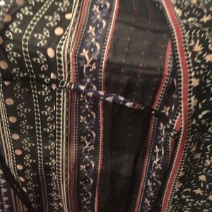 Op de afbeelding: Close-up van een patroonstof met verticale strepen en bloemmotieven. De stof heeft een mix van zwarte, witte, rode en blauwe kleuren. Het ontwerp bevat bloemen- en bladerpatronen, waardoor een bohemien esthetiek ontstaat.