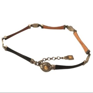 Op de afbeelding: Een decoratieve riem met afwisselende secties van bruin, zwart en bruin leer. De riem is geaccentueerd met zilverkleurige metalen hardware en een ketting sluiting. De centrale gesp heeft een goudkleurig medaillon.