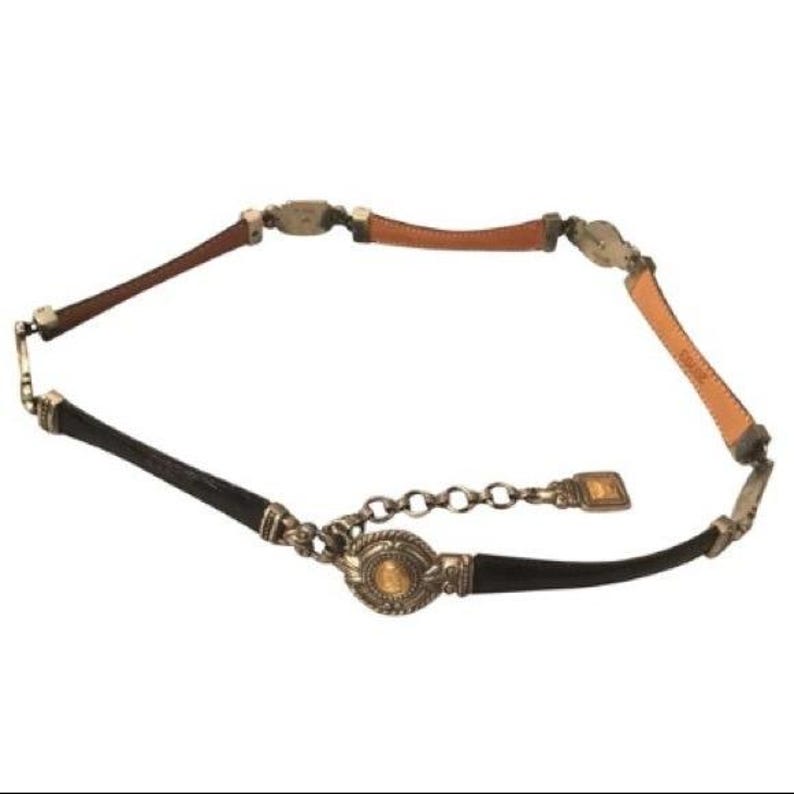 Op de afbeelding: Een decoratieve riem met afwisselende secties van bruin, zwart en bruin leer. De riem is geaccentueerd met zilverkleurige metalen hardware en een ketting sluiting. De centrale gesp heeft een goudkleurig medaillon.