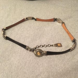 Op de afbeelding: Een decoratieve riem met bruine en zwarte leren secties, verbonden door zilverkleurige metalen hardware. De riem heeft een centraal medaillon met een goudkleurig centrum en een kettingverlenging voor verstelbare maatvoering.