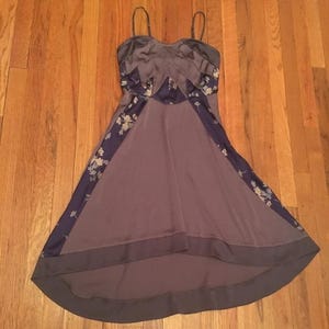 Puede incluir: Vestido sin mangas, largo hasta el suelo, en tonos grises y morados. El corpiño presenta un diseño geométrico y la falda tiene un dobladillo alto-bajo. Paneles azul oscuro con motivos florales recorren los laterales del vestido.