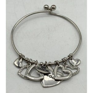 May include: Silver-toned bangle bracelet with heart charms. The bracelet features multiple heart-shaped charms, some with rhinestone accents, and a charm that reads "BBB Charming".