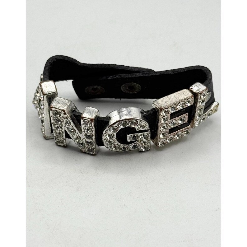 May include: A silver-toned bracelet spelling out "ANGEL" with clear rhinestones. The letters are mounted on a black leather band with snap closures. The bracelet is a fashion accessory.