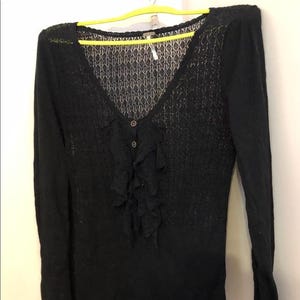 Pu&ograve; includere: Maglione nero a maniche lunghe con scollo a V e un delicato motivo a pizzo sulla parte superiore del petto. Il maglione presenta un dettaglio a balze sul davanti ed &egrave; allacciato con due bottoni. L'indumento &egrave; appeso a una gruccia gialla.